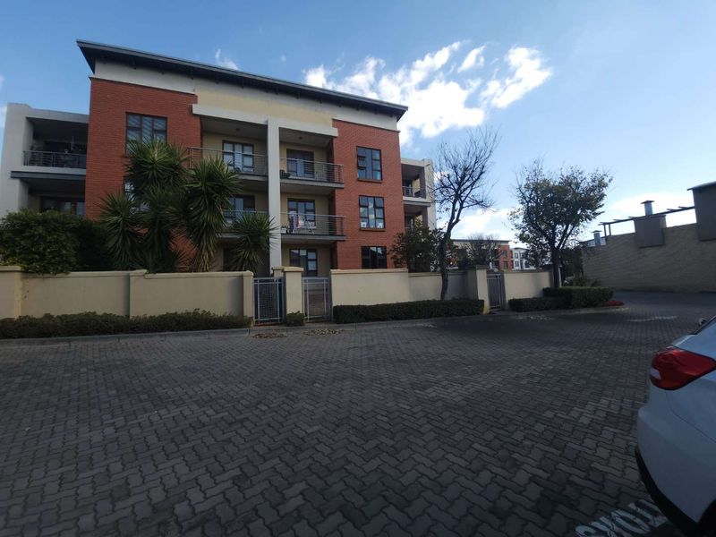 Exterior view of a modern multi-story apartment building featuring a brick facade, private balconies with metal railings, and a paved driveway entrance.