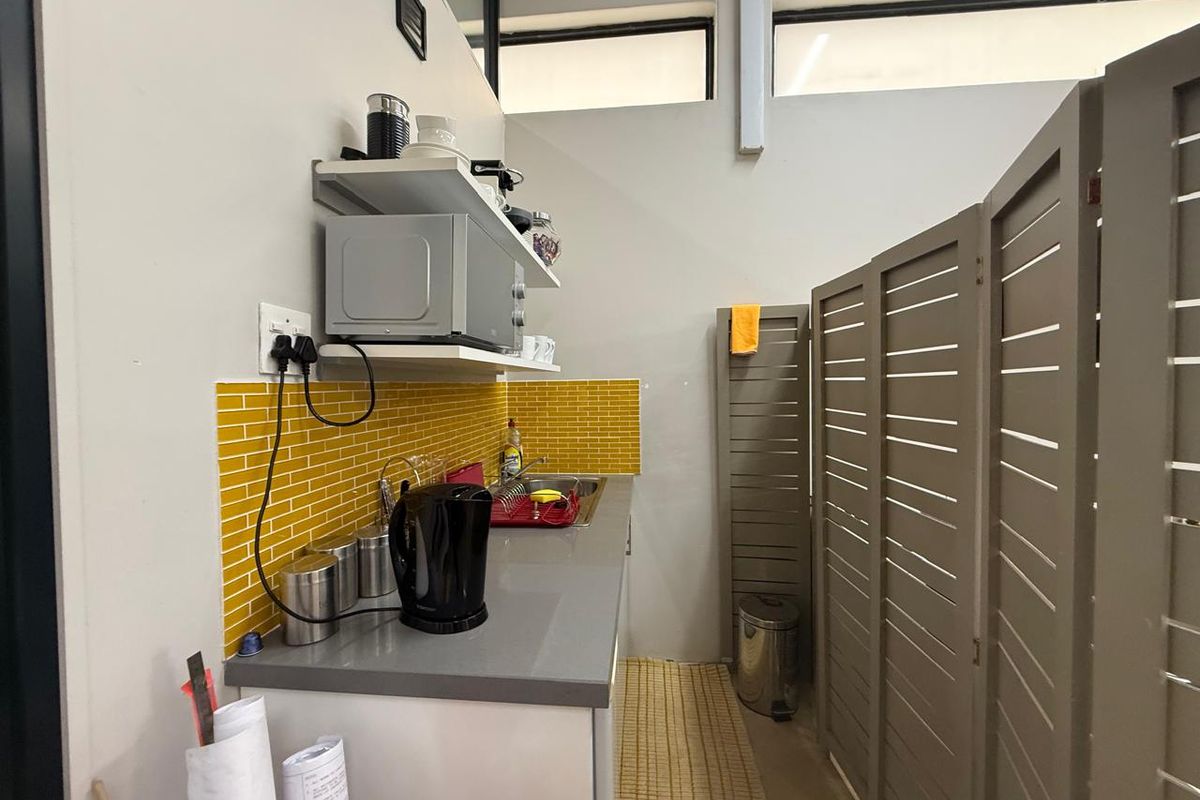 A compact office kitchenette featuring a grey countertop, stainless steel sink, and vibrant yellow subway tile backsplash with overhead shelving.