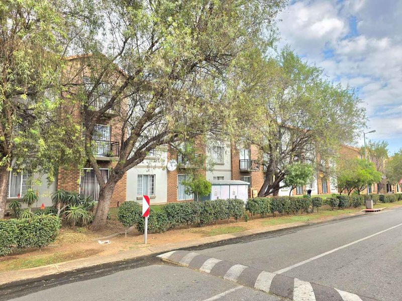 Street view of a multi-story apartment complex featuring brick facade construction, private balconies, and established trees along the roadside.