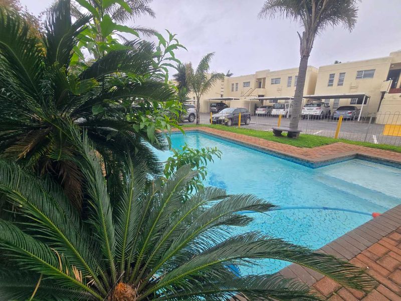 Communal swimming pool area featuring brick paving, perimeter fencing, and adjacent covered parking bays within the apartment complex.
