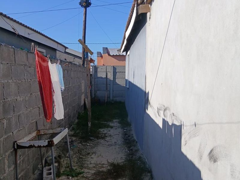 A narrow side passage between a concrete block boundary wall and the main building structure with an unpaved ground surface.