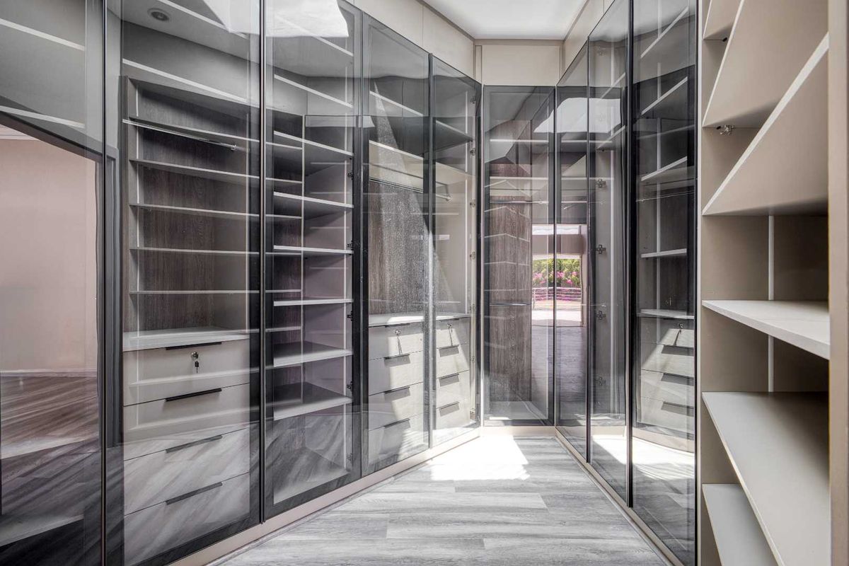 A modern walk-in closet featuring floor-to-ceiling glass-fronted cabinetry with integrated shelving and drawers, finished in a contemporary neutral palette.