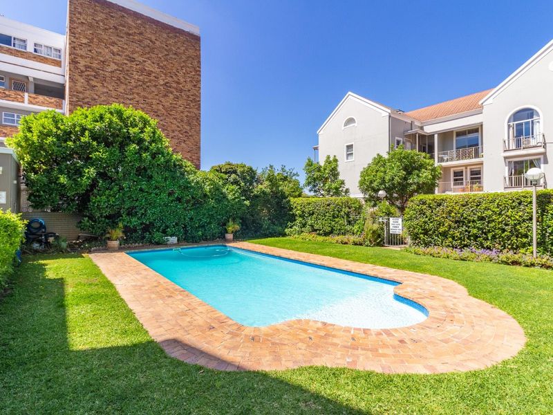 A well-maintained communal swimming pool area featuring brick coping, lush green lawns, and established garden hedging within an apartment complex setting.