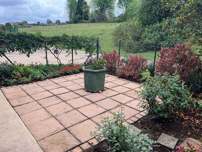 A paved outdoor patio area featuring established garden beds and perimeter chain-link fencing, overlooking a grassy landscape.