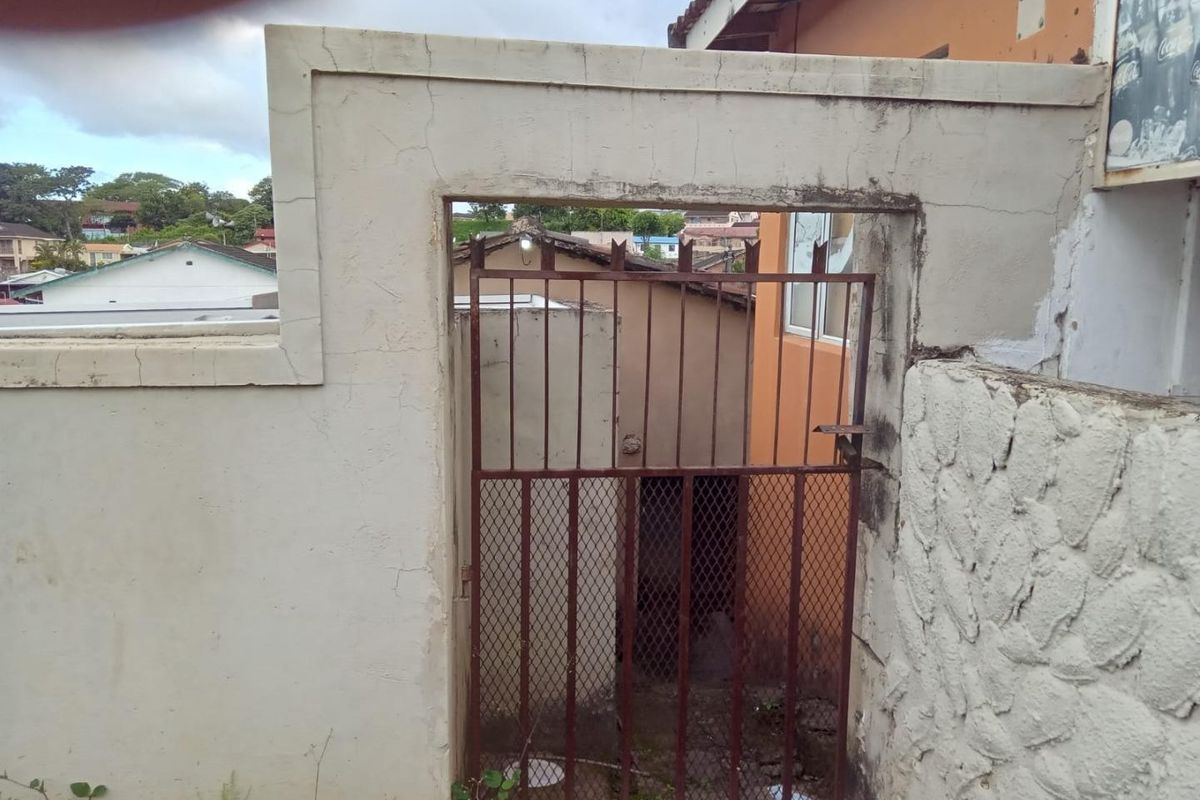 A rear exterior view showing a weathered masonry boundary wall with stone cladding and a rusted metal security gate leading to the property.