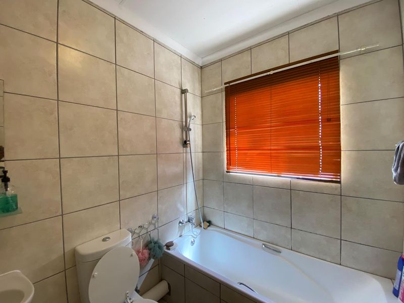 A functional bathroom featuring a bathtub with a shower mixer, a toilet, and neutral ceramic wall tiling with a window fitted with venetian blinds.