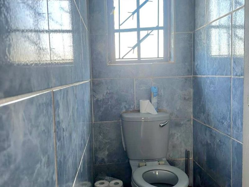 A small bathroom featuring blue ceramic wall and floor tiles, a standard toilet, and a window with security bars.