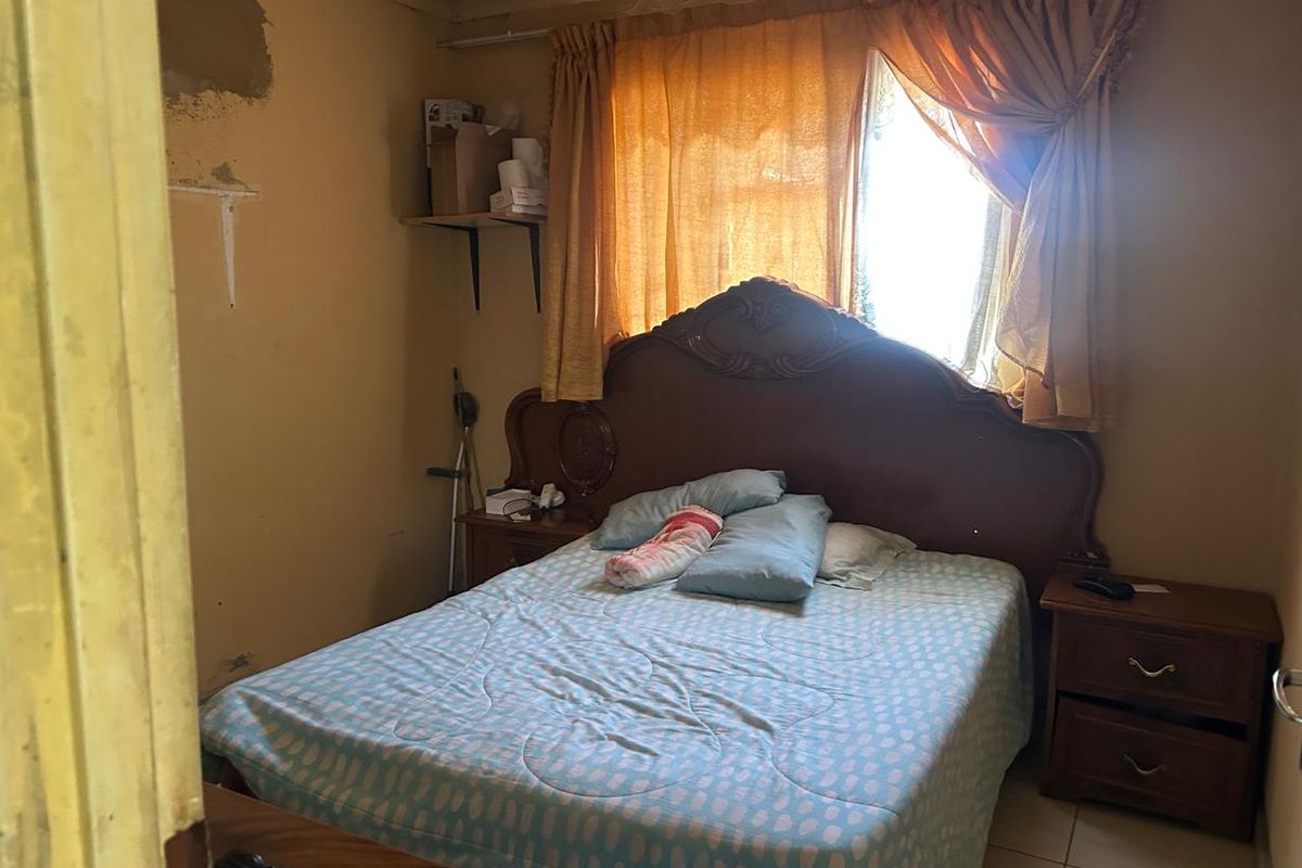 A bedroom featuring tiled flooring and a window with curtains, currently showing significant wear to the wall finishes.