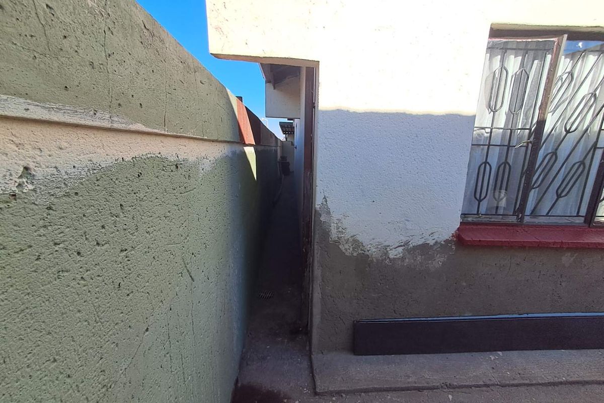 A narrow side passage between the exterior house wall and a boundary wall, featuring unfinished plasterwork and a concrete pathway.