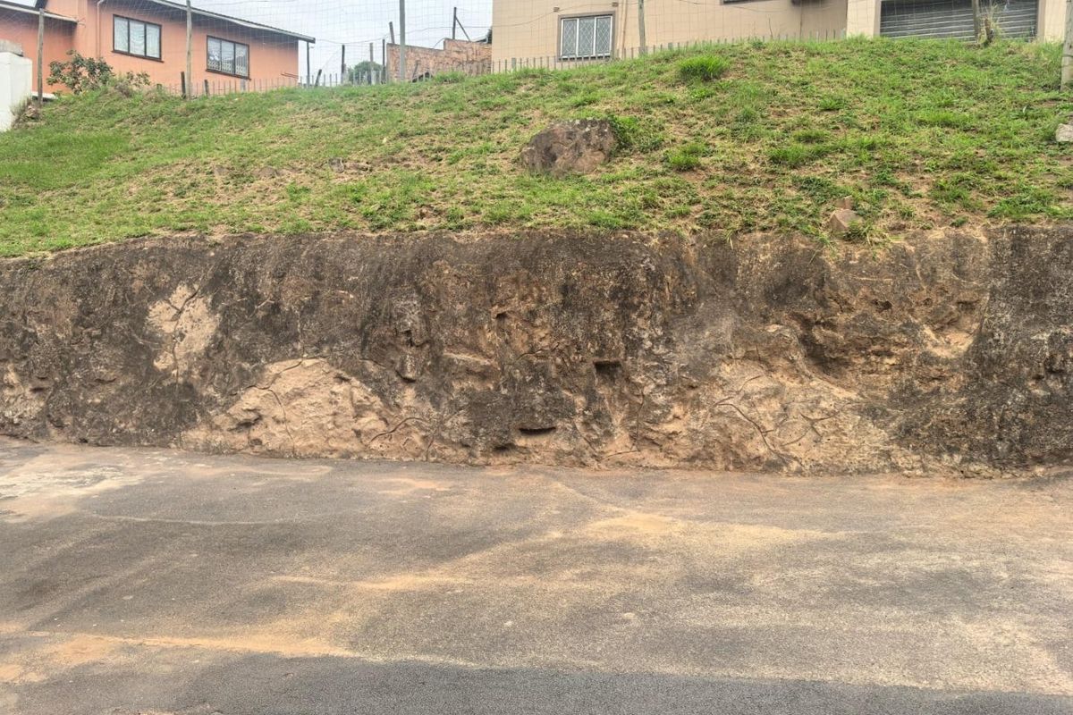 An exterior view of a property featuring a weathered, eroded retaining wall bordering an asphalt driveway and a grass embankment.