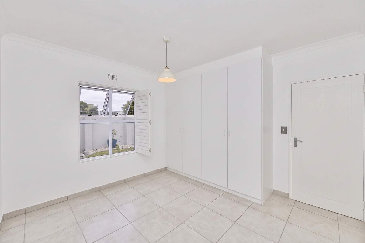 A bright, empty bedroom featuring neutral tiled flooring, built-in cupboards, and a window with shutter, offering a clean and versatile living space.