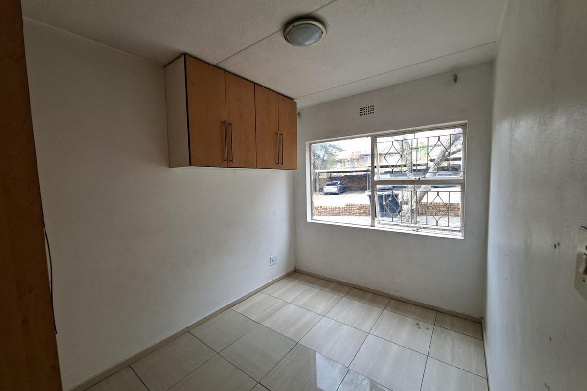 A compact bedroom featuring tiled flooring, a wall-mounted storage cabinet, and a window with security bars overlooking the exterior.