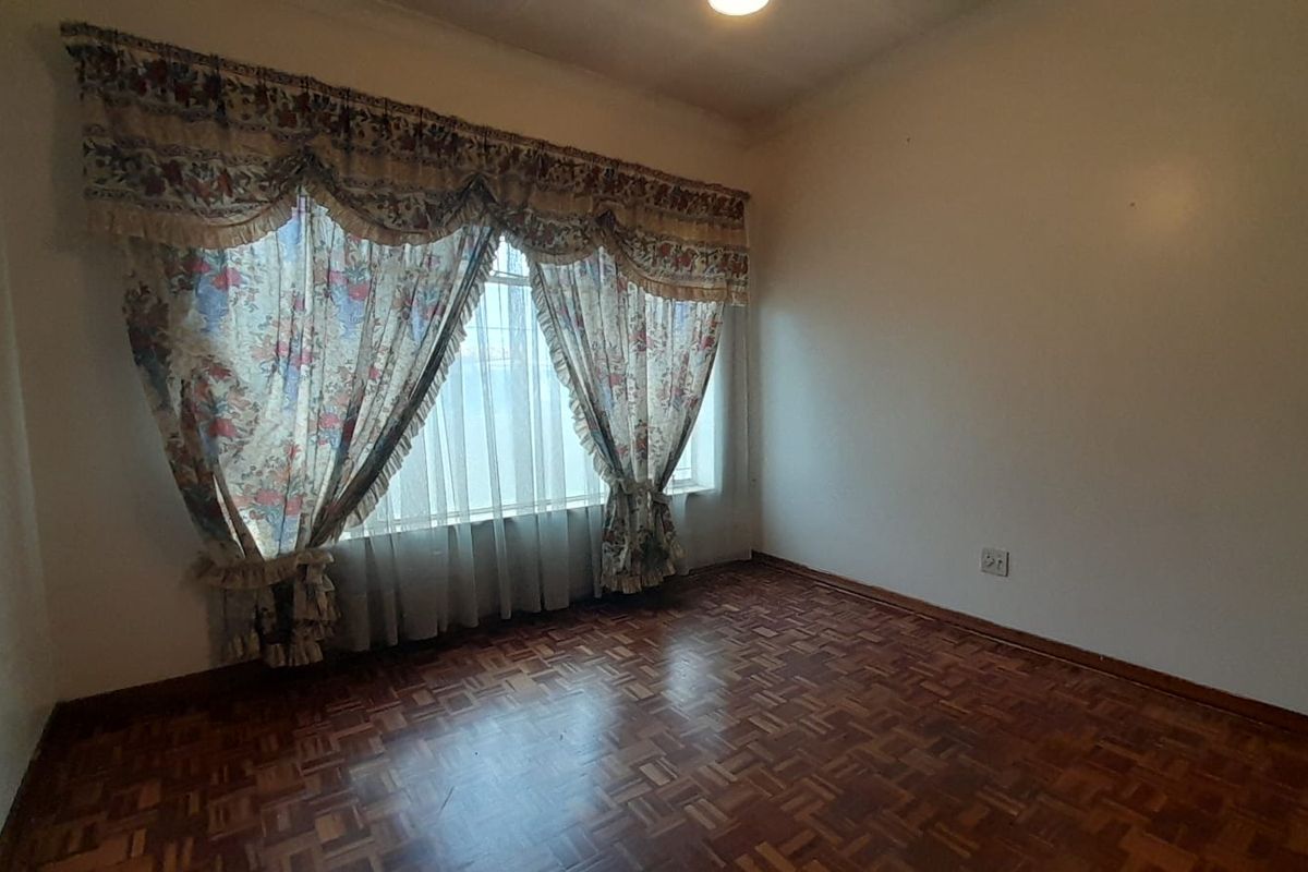 A vacant bedroom featuring classic parquet wood flooring and a large window with traditional curtain pelmets.
