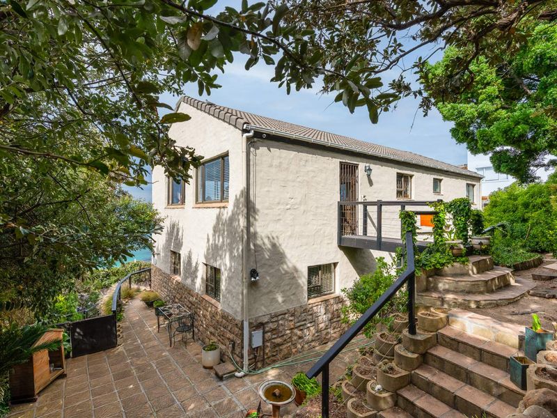 Rear view of the property featuring stone cladding, a raised balcony, and a stone-paved patio area surrounded by mature garden landscaping.