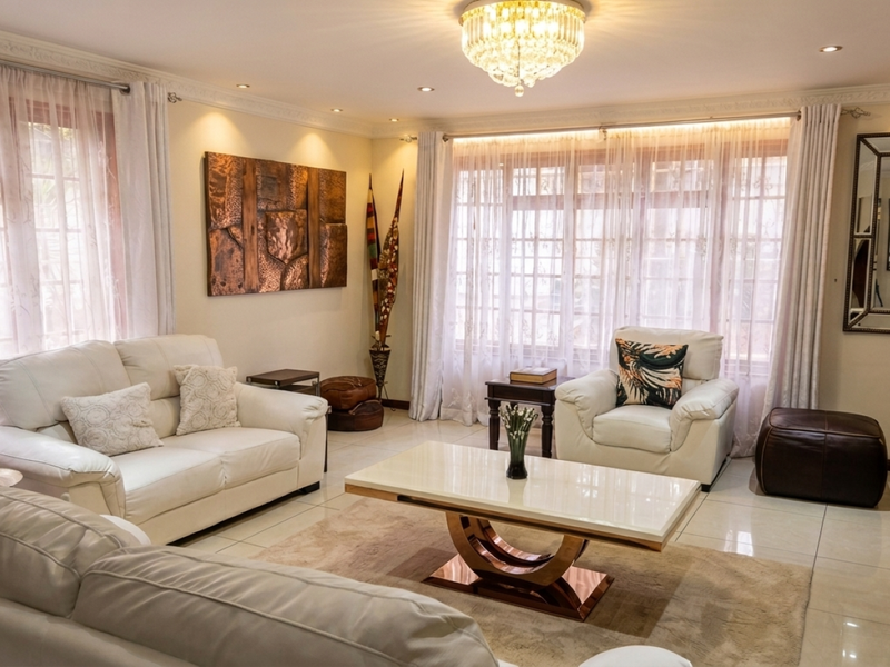 A well-maintained living room featuring tiled flooring, decorative ceiling cornices, recessed downlights, and a central crystal chandelier, providing a bright and inviting atmosphere.