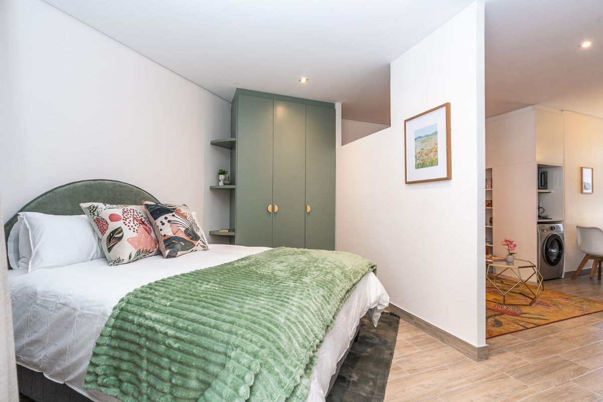 A modern bedroom featuring built-in sage green cabinetry, recessed lighting, and neutral tiled flooring, with an open-plan layout connecting to the adjacent living area.