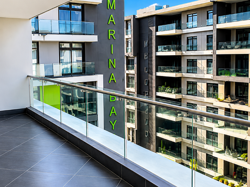 A modern, spacious balcony featuring sleek dark floor tiles and a contemporary glass balustrade, offering an elevated view of the surrounding apartment complex.