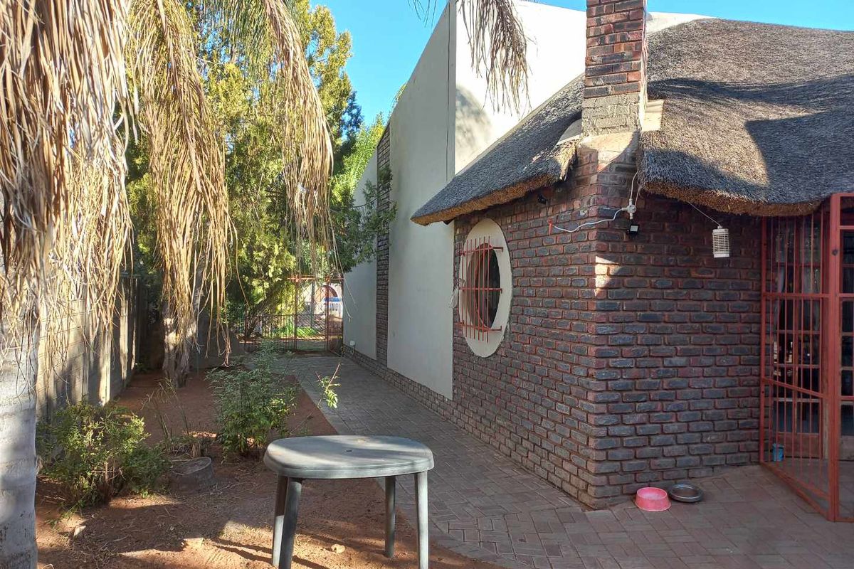 Rear exterior view of a house featuring a traditional thatch roof, exposed brickwork, a paved walkway, and a distinctive circular window with security bars.