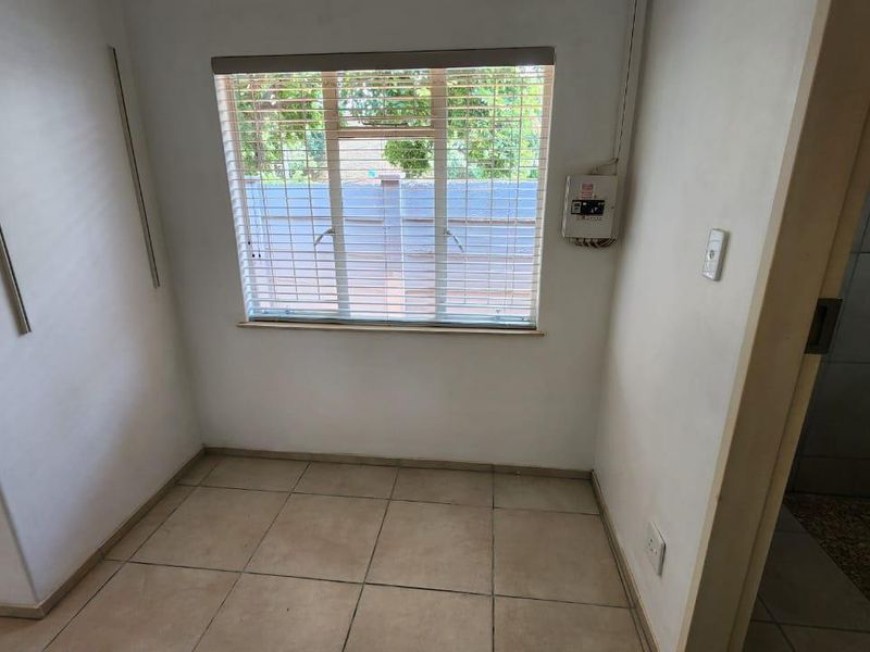 A compact, empty bedroom featuring tiled flooring, a window with venetian blinds, and an exposed electrical distribution board on the wall.