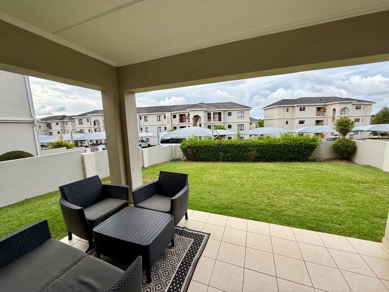 A covered patio area featuring tiled flooring, overlooking a private garden and the surrounding apartment complex.