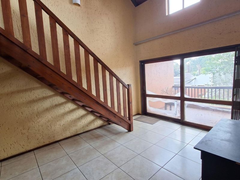 Spacious living area featuring a wooden staircase, high ceilings, tiled flooring, and large sliding glass doors leading to an outdoor balcony.