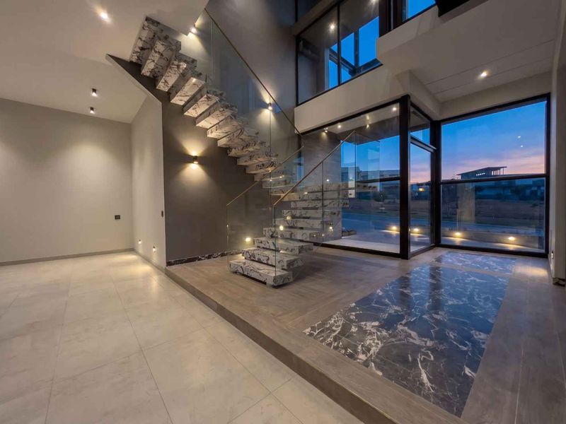 A modern, double-volume entrance hall featuring a striking marble staircase with glass balustrades, large format tiling, and expansive windows providing natural light.