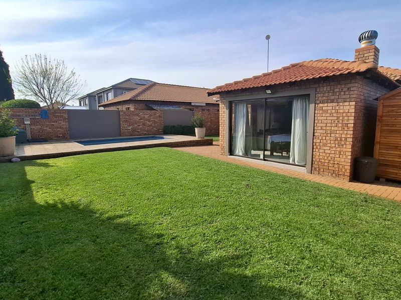A well-maintained rear garden featuring a manicured lawn, a swimming pool, a paved patio area, and a brick-built structure with sliding glass doors.