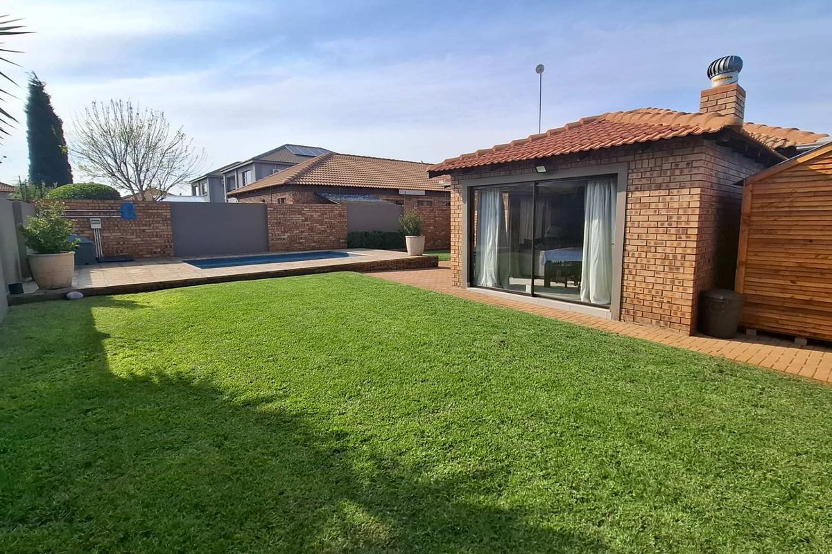 A well-maintained rear garden featuring a manicured lawn, a swimming pool, a paved patio area, and a brick-built structure with sliding glass doors.