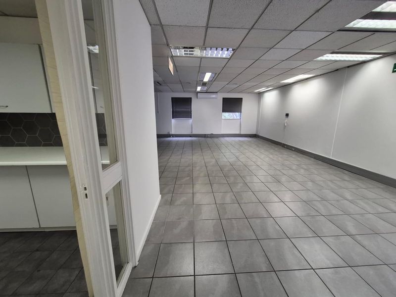 A spacious, empty open-plan office area featuring tiled flooring, a suspended ceiling with recessed lighting, and a small integrated kitchenette.