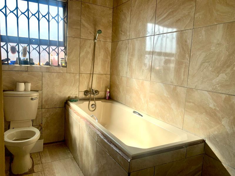A functional bathroom featuring a bathtub with a shower mixer, a toilet, and full-height wall tiling with security bars on the window.