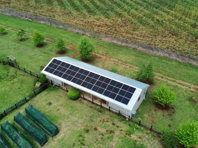 Aerial view of a farm outbuilding featuring a large roof-mounted solar panel array, surrounded by landscaped grounds and agricultural fencing.