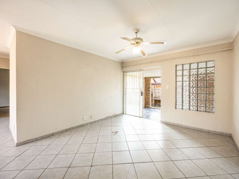 An empty living room featuring neutral tiled flooring, a ceiling fan, and a decorative glass block window with access to an outdoor patio area.