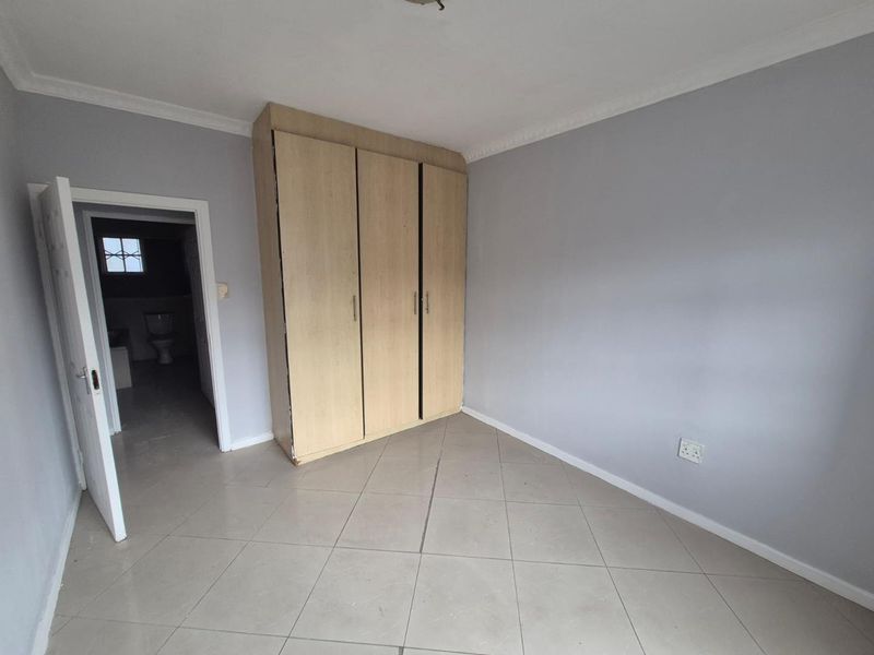 A clean, empty bedroom featuring neutral grey walls, tiled flooring, and a built-in wooden wardrobe, with an adjacent bathroom visible through the doorway.