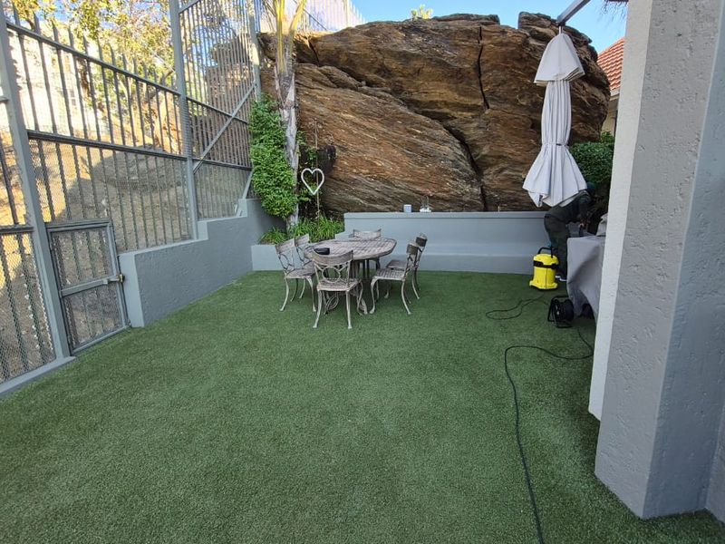 A private outdoor patio area featuring artificial turf, built-in bench seating, and a unique natural rock backdrop, enclosed by metal security fencing.