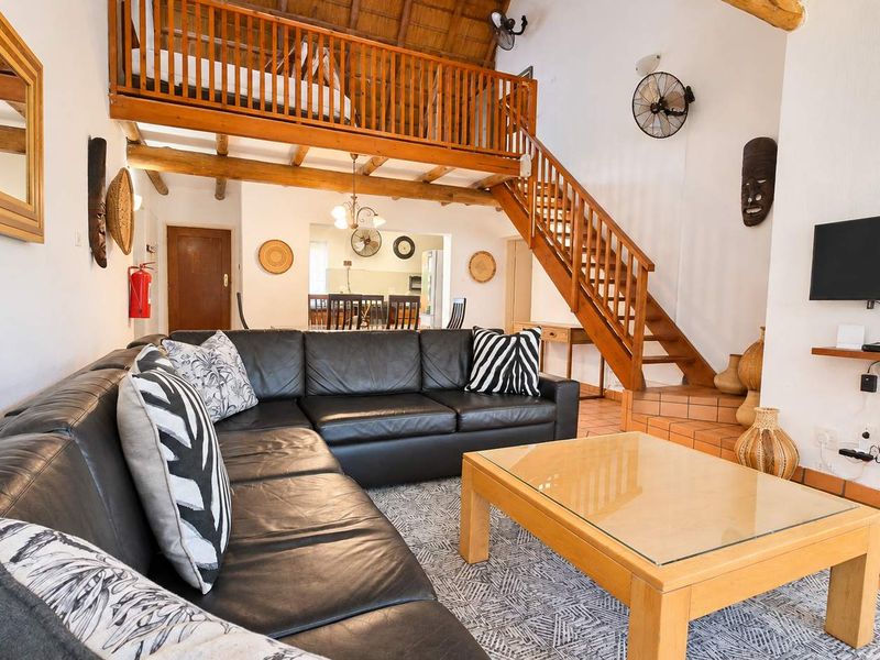 Open-plan living area featuring a mezzanine level, exposed timber beams, a thatched roof, and a wooden staircase, creating a spacious and rustic atmosphere.
