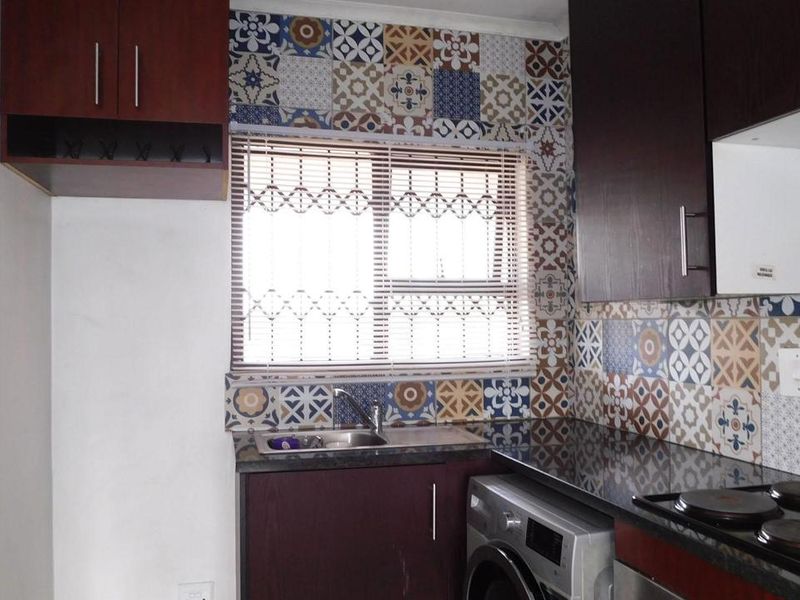 A compact kitchen featuring dark wood cabinetry, a patterned tiled splashback, integrated electric hob, and a stainless steel sink with a window view.