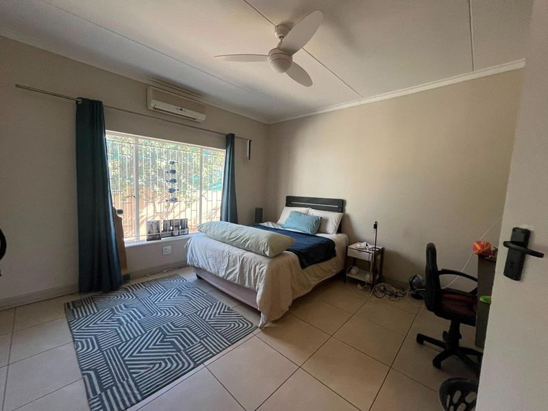 A functional bedroom featuring tiled flooring, a ceiling fan, and a wall-mounted air conditioning unit with security bars on the window.