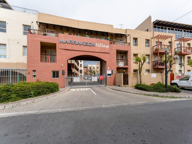 Front elevation of Marrakeh Village apartment complex featuring a secure gated entrance, paved driveway, and multi-level residential balconies.