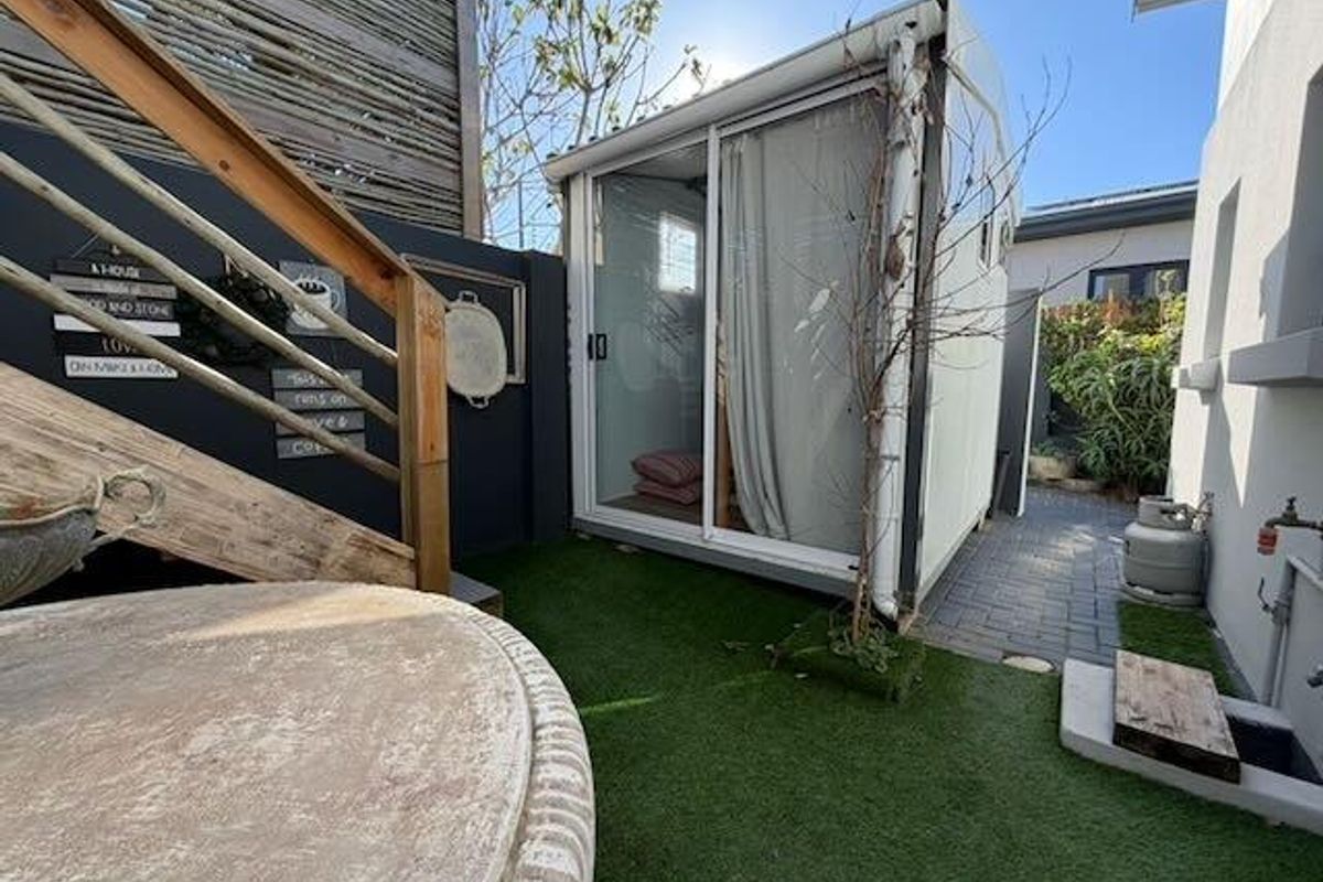 A compact outdoor area featuring a small detached studio or garden room with sliding glass doors, artificial lawn, and a wooden staircase.