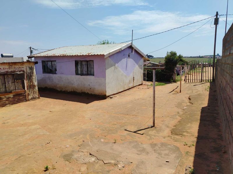 Rear exterior view of a modest house with a corrugated metal roof, security bars on windows, and a large unpaved yard area.
