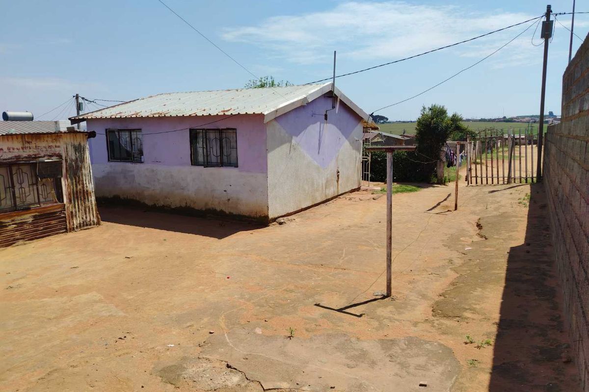 Rear exterior view of a modest house with a corrugated metal roof, security bars on windows, and a large unpaved yard area.