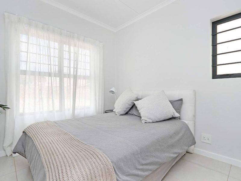 A bright and neutral bedroom featuring tiled flooring, a large window with sheer curtains, and a secondary high-level window.