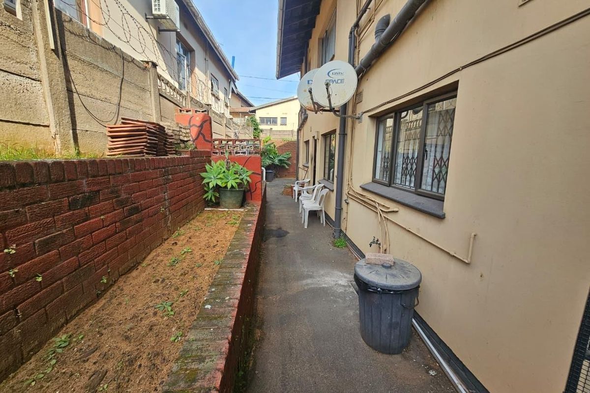 A narrow rear exterior walkway featuring a brick retaining wall, concrete path, and security-barred windows along the building facade.