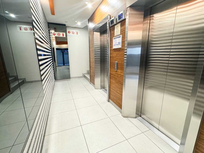 Modern office building elevator lobby featuring stainless steel lift doors, wood veneer wall paneling, and clean tiled flooring.