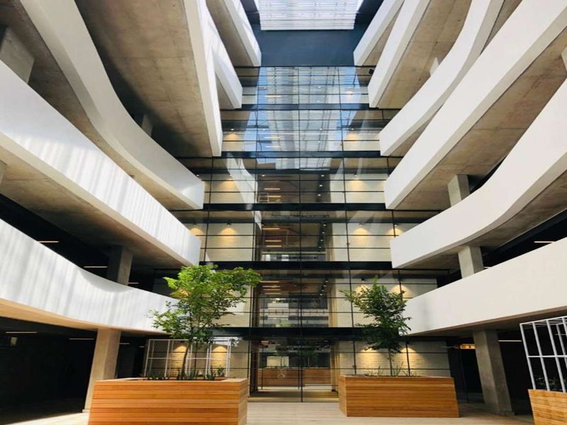 Modern architectural atrium featuring a central glass curtain wall, multi-level concrete balconies, and a large overhead skylight providing natural illumination.