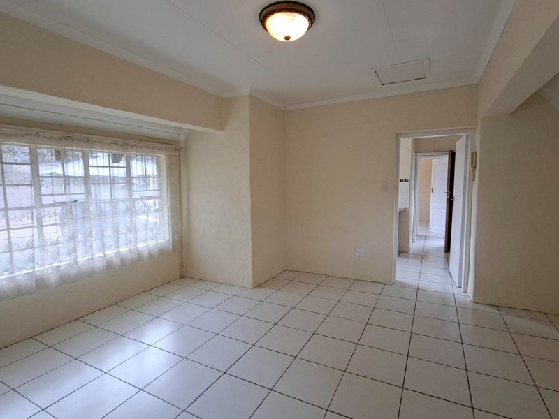 A bright, empty living area featuring neutral tiled flooring, a large window, and direct access to an adjoining hallway.