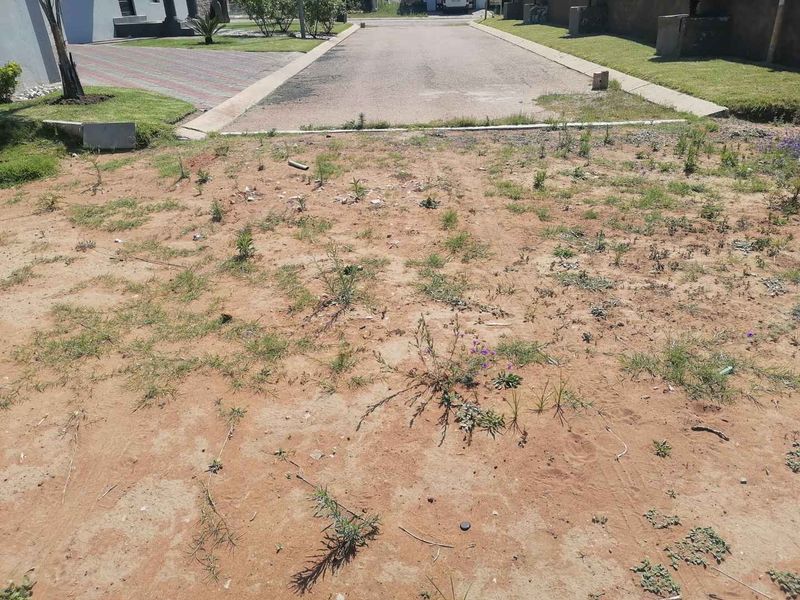 A view of a vacant residential plot showing the sandy ground surface and adjacent paved driveway and grass verge.