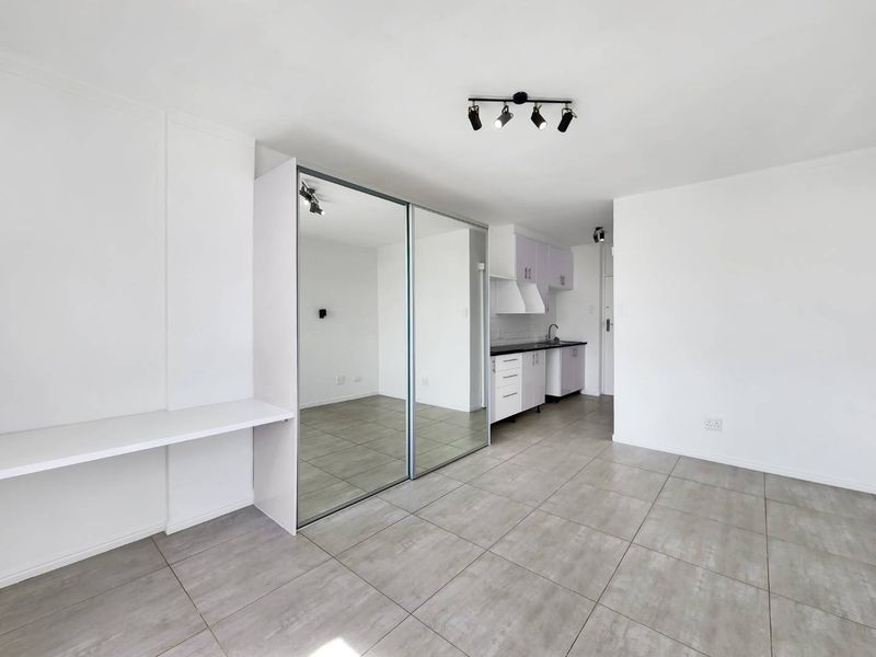 An open-plan living area featuring tiled floors, a built-in desk, mirrored sliding cupboards, and a compact kitchen with modern cabinetry.