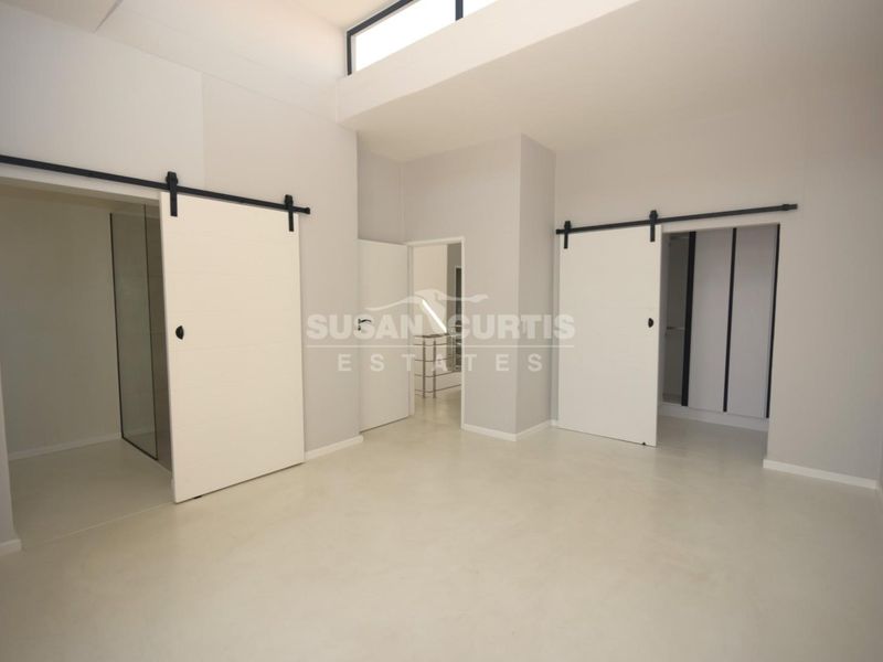 A modern, minimalist bedroom featuring polished concrete floors, high ceilings with clerestory windows, and dual sliding barn doors leading to an en-suite and wardrobe.