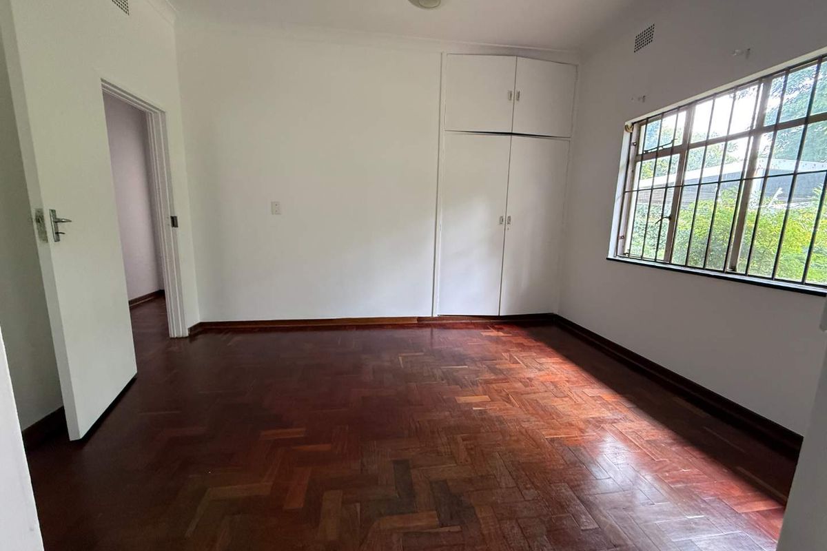 A bright, empty bedroom featuring classic parquet timber flooring, built-in storage cupboards, and security bars on the window.
